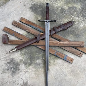 Könnte beinhalten: Ein langes, silbernes Schwert mit einem braunen, lederumwickelten Griff und einem Parier. Das Schwert wird mit seiner braunen Lederscheide präsentiert und liegt auf verwitterten Holzbrettern. Die Spitze der Scheide hat eine goldfarbene Metallspitze.