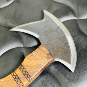 Peut inclure: Une hache à main avec une lame en acier poli et un manche en bois. Le manche est brun clair avec des motifs gravés et de petits rivets métalliques. La tête de hache a une lame incurvée et un sommet pointu.