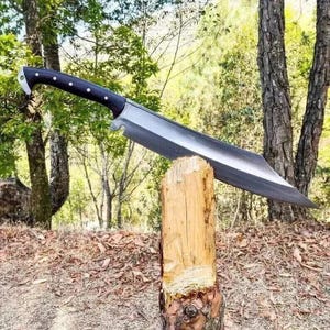 Puede incluir: Un cuchillo grande, hecho a mano, con un mango de madera oscura y una hoja ancha y plateada. El cuchillo descansa sobre un poste de madera en un entorno natural al aire libre.