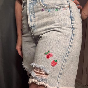以下が含まれることがあります： ハイウエストのアシッドウォッシュデニムショートパンツ。裾はフリンジ加工で、太もも部分にダメージ加工の破れがあり、サクランボとイチゴの刺繍ワッペンが付いています。フロントポケット付近には、小さくカラフルな多角形の宝石風の装飾が施されています。