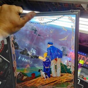 Puede incluir: Obra de arte náutica enmarcada que representa a un marinero y un niño mirando al mar, con un perro pequeño. La obra presenta una combinación de colores azules y blancos, con un borde en forma de cuerda. La escena incluye gaviotas, un faro y una casita.