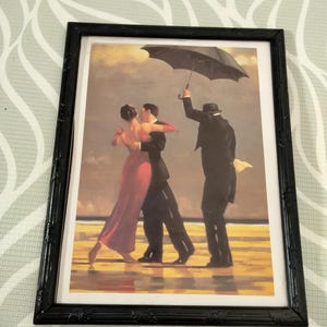 Puede incluir: Impresión artística enmarcada que representa a una pareja bailando en una playa, con un hombre sosteniendo un paraguas. La mujer lleva un vestido rojo y el hombre un traje negro. El marco es negro.