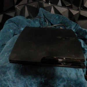 May include: A black PlayStation 3 game console featuring a sleek, rectangular design with a glossy finish. The console has the PlayStation logo embossed on the top surface and a disc slot located on the front panel, with a power cable extending from the back.
