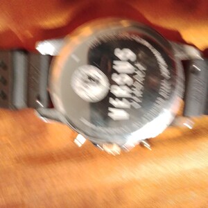 May include: A silver-toned Versus Versace watch with a black rubber strap. The watch face is visible, with the brand name and logo. The watch is set against an orange background.
