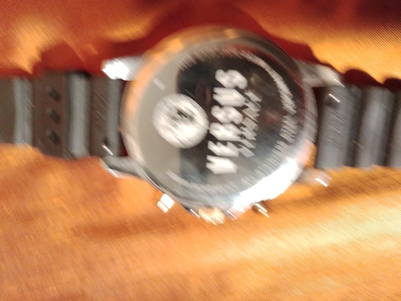May include: A silver-toned Versus Versace watch with a black rubber strap. The watch face is visible, with the brand name and logo. The watch is set against an orange background.
