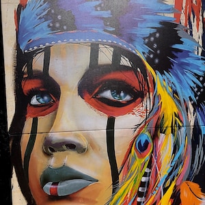 Puede incluir: Un lienzo impreso que muestra el retrato estilizado de una persona con ojos azules, que lleva un tocado de plumas azules y pintura facial. La obra incorpora plumas amarillas, naranjas y azules vibrantes, con líneas negras verticales en la cara.