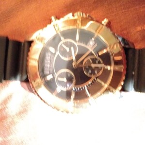 May include: A gold-toned wristwatch with a black face and three sub-dials. The watch has a black rubber strap. The watch face has gold-toned hands and markers. The watch is a luxury item.
