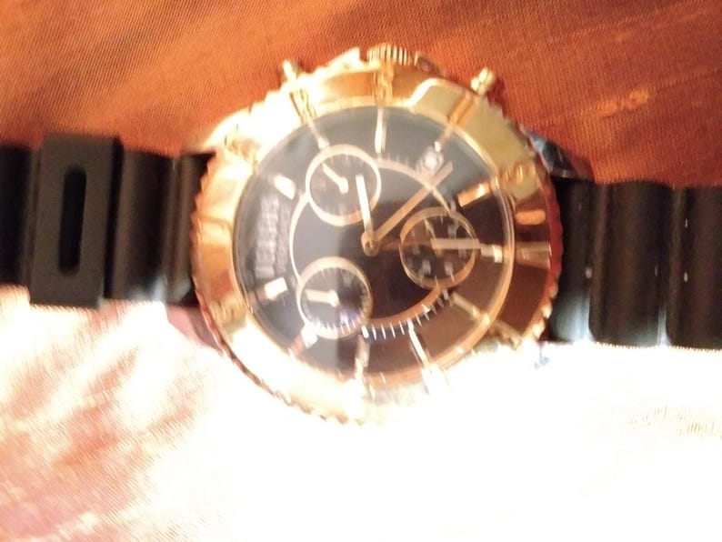 May include: A gold-toned wristwatch with a black face and three sub-dials. The watch has a black rubber strap. The watch face has gold-toned hands and markers. The watch is a luxury item.
