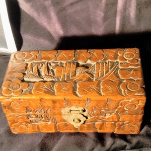 Può includere: Una scatola rettangolare in legno con finitura marrone medio, caratterizzata da intricati motivi floreali intagliati a mano lungo i bordi e una scena paesaggistica centrale sul coperchio. La scatola include una chiusura in metallo color ottone.