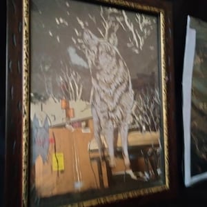 Puede incluir: Una pieza de arte mural enmarcada que presenta una ilustración de un lobo blanco sobre un fondo de bosque oscuro y nevado. La obra está encerrada en un marco de madera marrón oscuro con detalles dorados, mostrando una escena invernal con árboles.