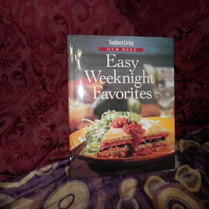 Puede incluir: Un libro de cocina de Southern Living titulado Our Best Easy Weeknight Favorites. La portada muestra una fotografía de un plato en capas sobre un plato naranja, cubierto con salsa roja y guarnición verde, con un tenedor al lado.