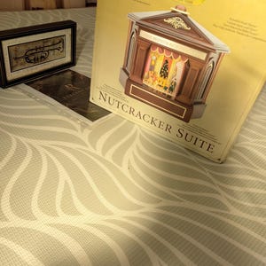 May include: A brown and gold "Nutcracker Suite" musical theater box with a detailed stage scene. A framed trumpet art piece and a brochure are also visible. The box is from the Gold Label Collection.