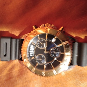 May include: A gold-toned Versus Versace watch with a black dial and three sub-dials. The watch has a black rubber strap and a gold bezel. The watch is set against a textured orange background.