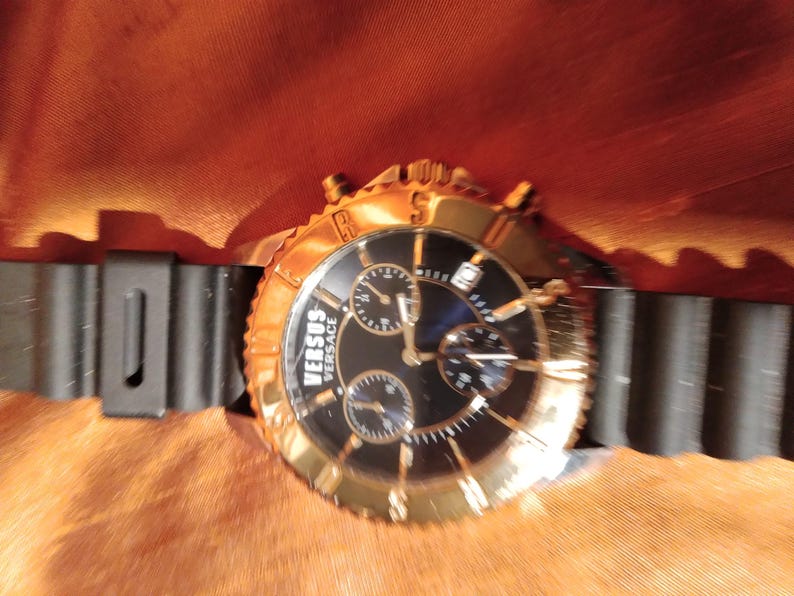 May include: A gold-toned Versus Versace watch with a black dial and three sub-dials. The watch has a black rubber strap and a gold bezel. The watch is set against a textured orange background.