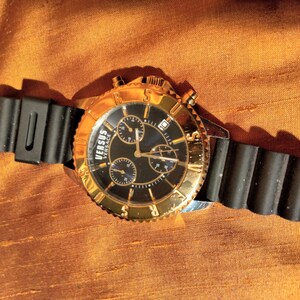 May include: A gold-toned Versus Versace watch with a black dial and three sub-dials. The watch features a gold bezel and a black rubber strap. The watch is displayed on a textured, orange-brown surface.