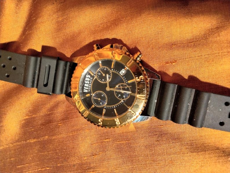 May include: A gold-toned Versus Versace watch with a black dial and three sub-dials. The watch features a gold bezel and a black rubber strap. The watch is displayed on a textured, orange-brown surface.