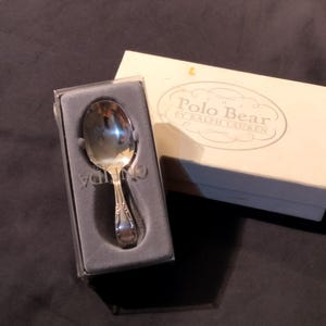 Puede incluir: Una cuchara plateada en una caja forrada de terciopelo gris. La cuchara tiene un mango decorativo y está empaquetada con una caja blanca que dice "Polo Bear by Ralph Lauren". Es un utensilio de cocina.
