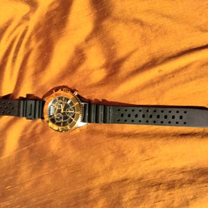 May include: A gold-toned wristwatch with a black dial and a black rubber strap. The watch features a gold bezel and a chronograph design. The watch is lying on a textured, orange-yellow surface.