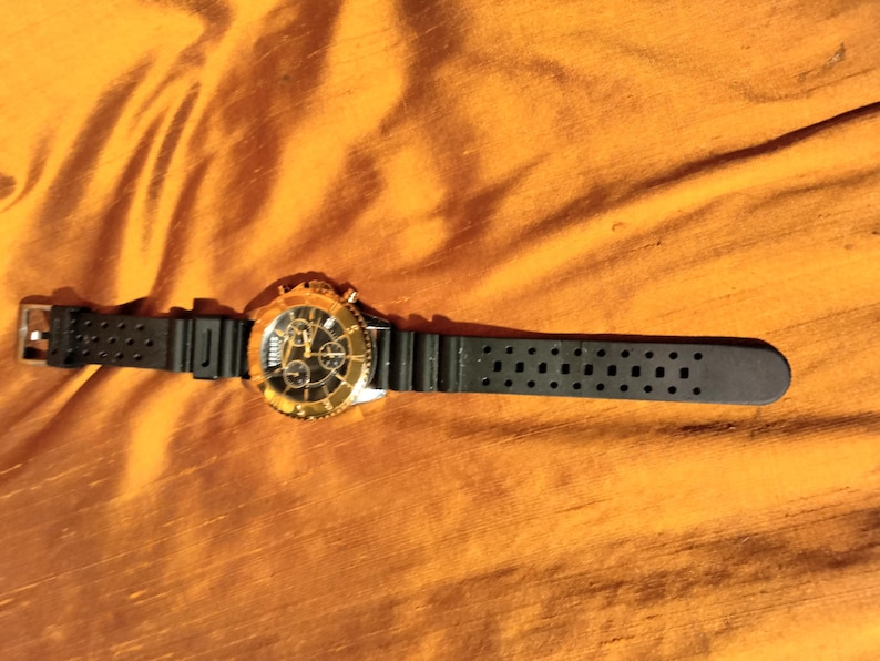 May include: A gold-toned wristwatch with a black dial and a black rubber strap. The watch features a gold bezel and a chronograph design. The watch is lying on a textured, orange-yellow surface.