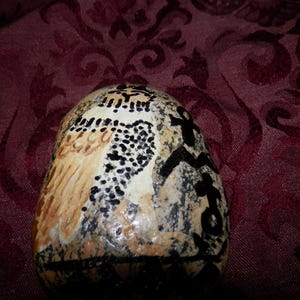 以下が含まれることがあります： 茶色、クリーム色、黒の色合いで手描きされたフクロウのデザインが施された、滑らかで斑点のあるベージュとグレーの川石。フクロウは詳細な羽毛と大きな目で描かれており、側面にはアンクに似た黒いインクのシンボルが添えられています。