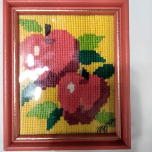 Puede incluir: Arte de tapiz enmarcado con dos manzanas rojas con hojas verdes sobre un fondo amarillo. La obra de arte está encerrada en un marco rectangular de madera de color coral con un borde dorado. Las manzanas tienen reflejos blancos.
