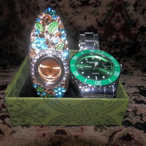 May include: A decorative watch with a floral-patterned gold-tone frame adorned with blue and green enamel leaves and clear crystals, alongside a silver-tone watch with a green bezel and a green dial, both presented in a green patterned box.