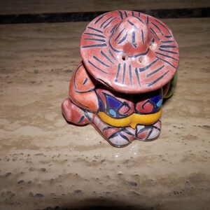 Puede incluir: Un salero y pimentero de cerámica con forma de figura que lleva un sombrero de ala ancha. La figura está pintada en tonos terracota, amarillo y azul con detalles de líneas negras. El sombrero tiene pequeños orificios para el condimento.