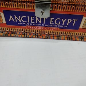 Puede incluir: Un cofre del tesoro rectangular con un cierre metálico. El cofre presenta un diseño con jeroglíficos egipcios y las palabras "ANCIENT EGYPT" en letras grandes sobre un fondo azul. El texto dice "3.000 AÑOS DE MISTERIO PARA DESBLOQUEAR Y DESCUBRIR COFRES DEL TESORO."