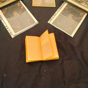 Puede incluir: Un libro abierto con páginas naranjas está centrado en una superficie negra, rodeado de arte enmarcado y documentos. Los documentos tienen la palabra "RELIANCABAS" impresa. Las obras de arte tienen marcos dorados.