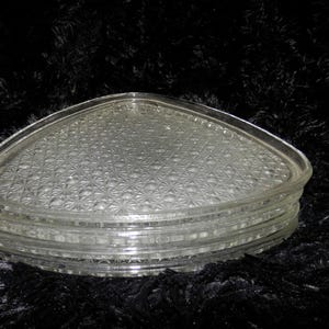 May include: A stack of three clear glass plates with a triangular shape and rounded corners. Each plate features a textured, pressed starburst or geometric pattern on the surface, with a smooth, raised rim around the outer edge of the glass.
