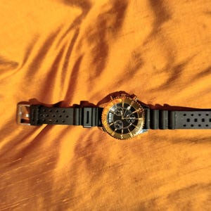 May include: A Versus watch with a black band and a gold-colored bezel and face. The watch has a black dial with gold-colored hands and markers. The watch is on an orange background.