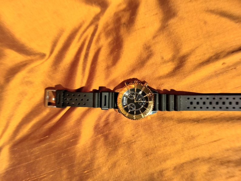 May include: A Versus watch with a black band and a gold-colored bezel and face. The watch has a black dial with gold-colored hands and markers. The watch is on an orange background.