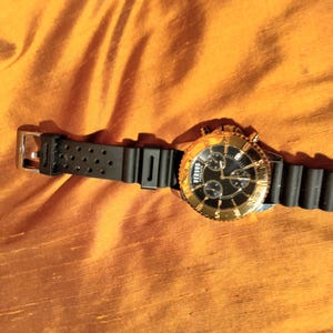 May include: A Versus gold-tone watch with a black dial and black rubber strap. The watch features a gold bezel and a black strap with multiple holes. The watch is displayed on an orange fabric surface.