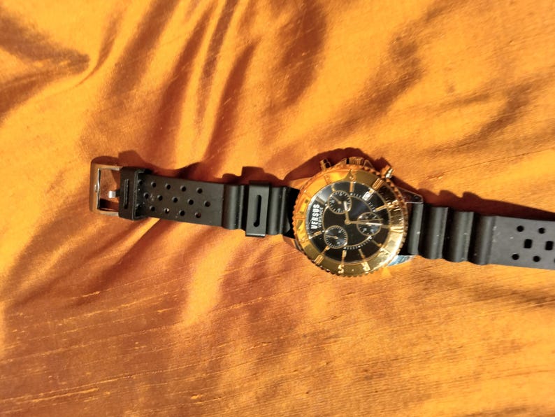May include: A Versus gold-tone watch with a black dial and black rubber strap. The watch features a gold bezel and a black strap with multiple holes. The watch is displayed on an orange fabric surface.