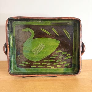 May include: A rectangular ceramic tray with rounded corners and two side handles. The interior features a dark brown background with a hand-painted green swan motif, accented by small green leaf-like shapes and a cross-hatched pattern on the body.