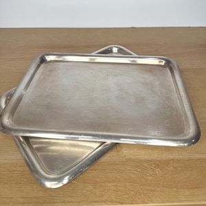 May include: Two rectangular silver-toned metal serving trays with rounded corners and raised edges. The trays have a smooth, reflective surface with a subtle metallic sheen and are stacked slightly offset from one another on a flat surface.