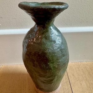 May include: A dark green ceramic vase with a textured, uneven surface and a wide, flared rim. The body of the vase is bulbous, tapering toward a narrow neck, and it features a visible, unglazed, light-colored base with a coarse, sandy texture.