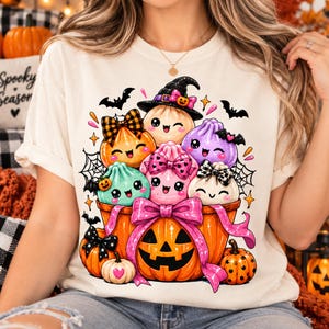 Peut inclure: Un t-shirt couleur crème arborant un motif de plusieurs personnages boulettes colorés et souriants avec des nœuds et des chapeaux, nichés dans une grande citrouille orange décorée d'un ruban et d'un nœud rose vif.
