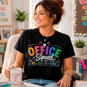 Può includere: Una t-shirt nera a maniche corte con il testo colorato "OFFICE Squad" e "WE GET IT DONE" accompagnato da una piccola icona a forma di cuore bianco. Il design presenta lettere color arcobaleno e dettagli bianchi sul davanti del capo.