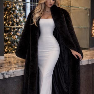 May include: A woman wearing a long, black faux fur coat over a white, strapless, floor-length dress. The coat is open, revealing the dress. The background includes a Christmas tree and a marble countertop.