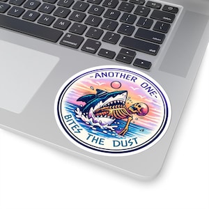 May include: A round sticker featuring a shark biting a skeleton in a sea scene with a sunset background. The sticker has the text "-ANOTHER ONE- BITES THE DUST". The sticker is placed on a laptop.