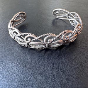 以下が含まれることがあります： 手書き風の「Welcome」の文字が入った、白く塗装された素朴な木製サイン。 傷んだ風合いで、装飾的な花の要素が特徴です。 背景は家のぼやけた画像です。