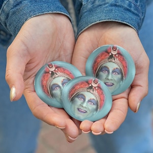 May include: Three round, light blue buttons featuring a smiling face with green skin, blue eye makeup, and a red and gold turban. The buttons are held in cupped hands against a denim background.