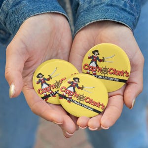 May include: Three round yellow pinback buttons featuring a cartoon pirate character holding two swords, a ship wheel graphic, and the red text Cap'n Clark's above the black text Ottoman Empire on a horizontal black stripe across the center.