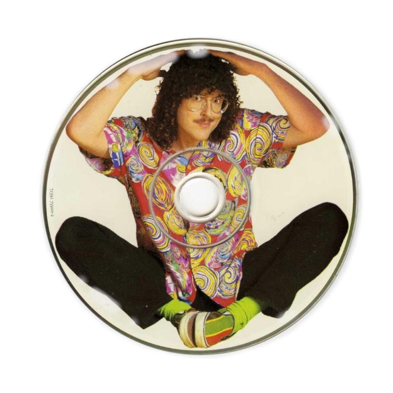 May include: A CD featuring a man with curly hair, glasses, and a colorful shirt. He is sitting with his legs crossed and arms raised, with the image of the man printed on the CD. The shirt has a swirl pattern in red, yellow, and blue.