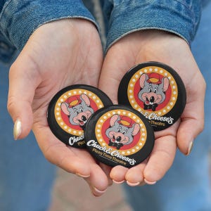 May include: Three round black buttons featuring the Chuck E. Cheese's Pizza Time Theatre logo. Each button has a cartoon mouse wearing a hat and bow tie, surrounded by a red and yellow design. The text "Chuck E. Cheese's Pizza Time Theatre" is also visible.