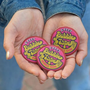May include: Three pink bubble gum packages are held in cupped hands. Each package has the text "6 FEET Bubble Tape Bubble Gum" in yellow and white. The packages are round and have a retro design.