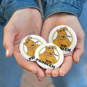 May include: Three white round buttons featuring the character ALF, with a yellow border. The buttons have the text "ALF!" and "NO PROBLEM" printed on them. The buttons are held in open hands.