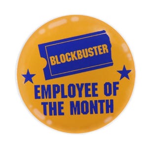 May include: A round, yellow pin with the words "BLOCKBUSTER EMPLOYEE OF THE MONTH" in blue. The pin features a blue rectangle with the word "BLOCKBUSTER" and two blue stars.