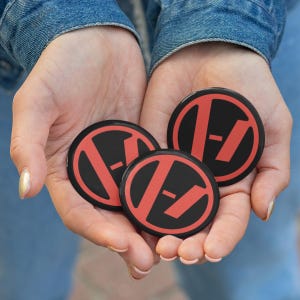 May include: Three black and red circular buttons are held in cupped hands. Each button features a red design within a black circle. The design includes a red slash and a horizontal line. The buttons are displayed against a denim background.